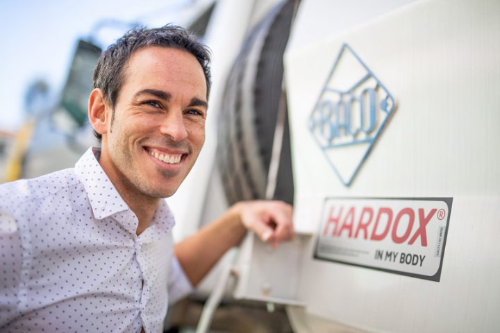 Начальник производства компании Industrias Baco с улыбкой на лице стоит рядом с автомобилем, отмеченным знаком качества «Hardox® In My Body».