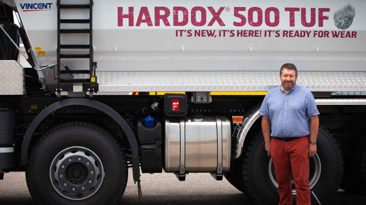 Hymyilevä työntekijä seisoo Hardox® 500 Tuf -teräksestä valmistetun kippilavan edessä.