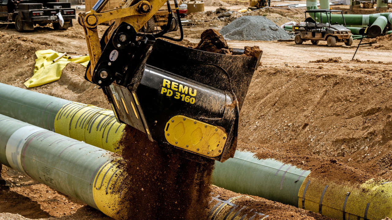 A screening bucket empties soil over pipework on a construction site