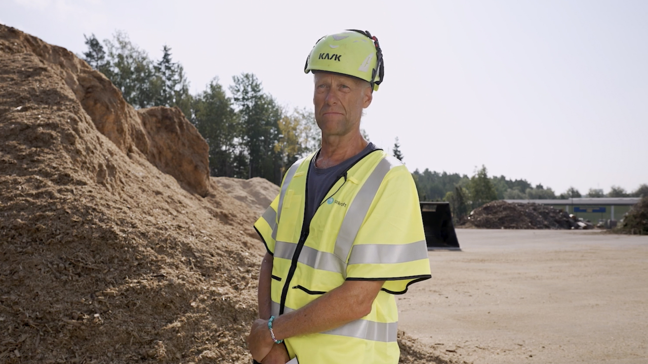 Un muncitor stând în fața unei stive uriașe de biomasă din așchii de lemn într-o centrală de termoficare.