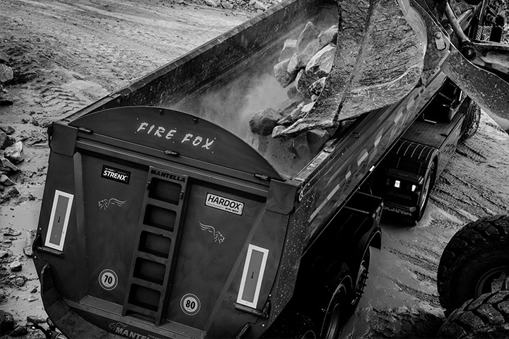 A bucket dumping out abrasive rocks into a Firefox truck body bearing the Hardox® In My Body logo.