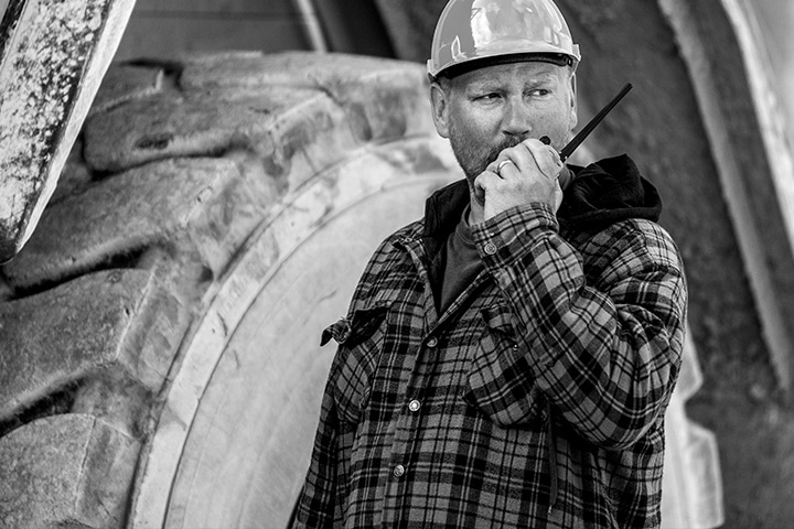 Worker in a hard hat using a walkie-talkie, standing next to a giant wheel from a mining haul truck.