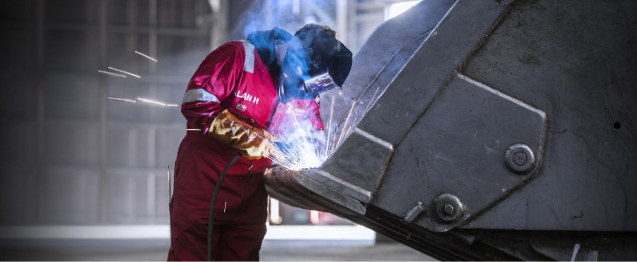 Egy védősisakos hegesztőmunkás nagy szilárdságú Hardox 400 acélból készült kopásálló acéllemezt hegeszt egy kotrógépkanál belsejében.