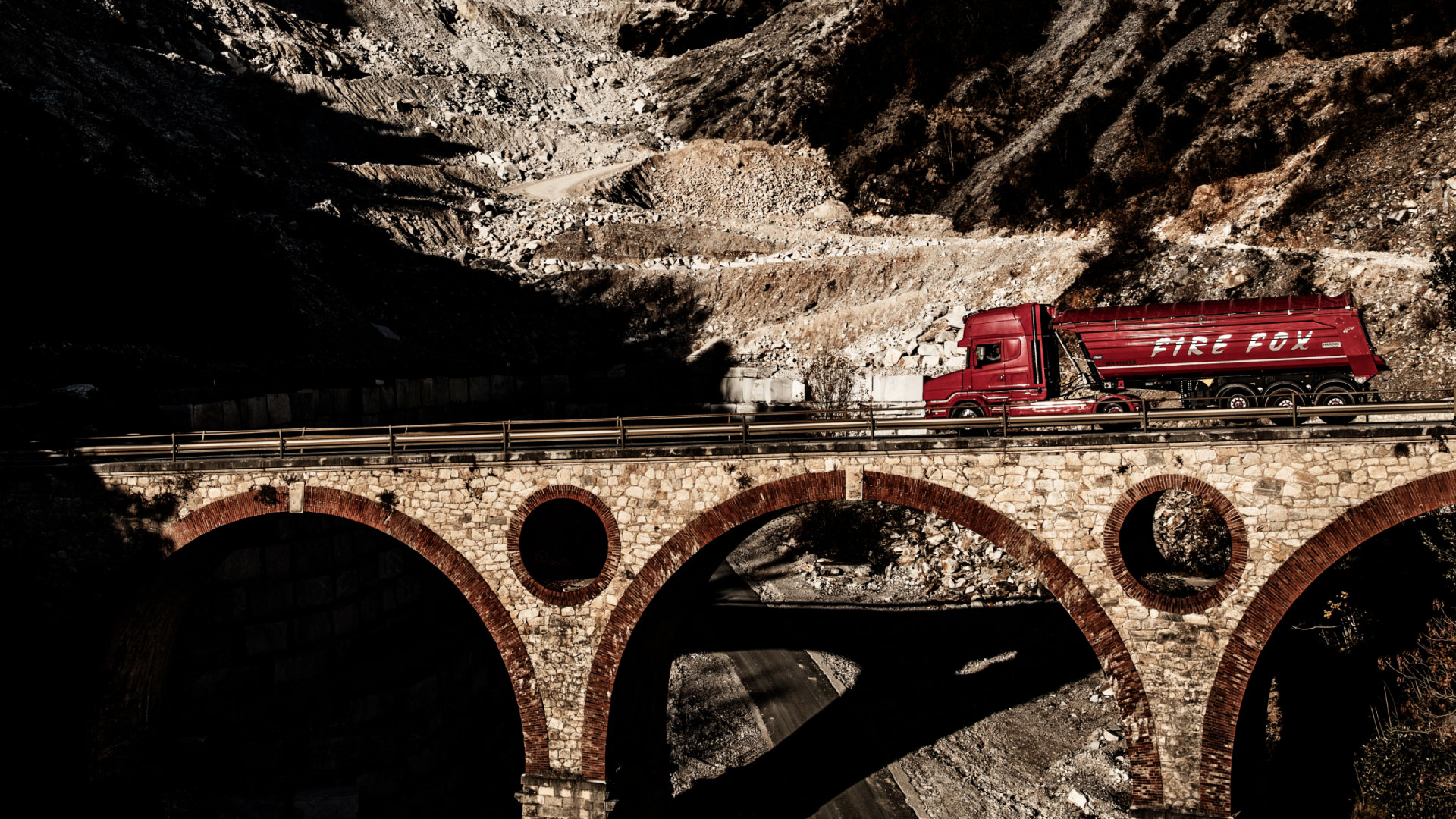 Chiếc xe tải màu đỏ, với thùng xe được thiết kế kiểu trọng lượng nhẹ bằng thép Hardox®, đang chạy qua cây cầu đá.